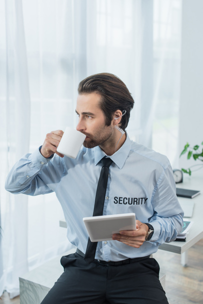 Ασφάλεια άνθρωπος στο ακουστικό κρατώντας φορητό υπολογιστή και πίνοντας τσάι στο γραφείο - Φωτογραφία, εικόνα