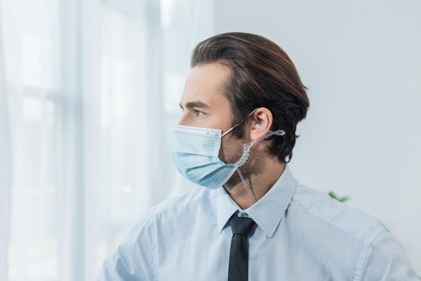 young security man in earphone and medical mask looking away in office - Foto, Imagem