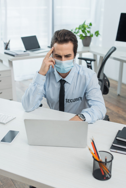 security man in medical mask looking at computer monitor near smartphone with blank screen in supervision room - Photo, Image