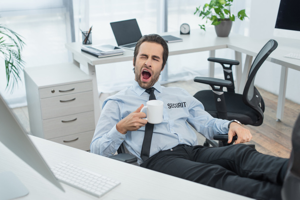 bored guard with cup of tea yawning while sitting with legs on desk in office - Photo, Image