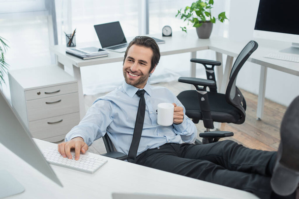 cheerful security man with cup of tea looking at monitor while sitting with legs on desk - Photo, Image