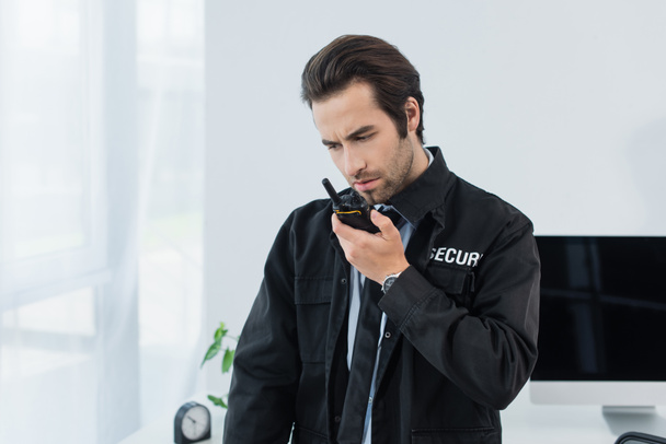 homme de sécurité sérieux en uniforme noir appelant walkie-talkie dans le bureau - Photo, image