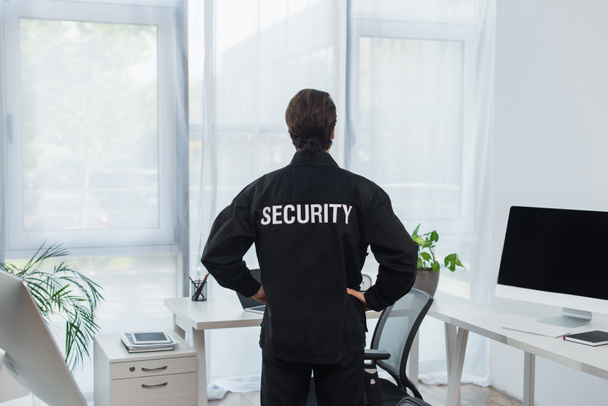 vista trasera de la guardia en uniforme negro con letras de seguridad de pie cerca de las computadoras en la oficina - Foto, Imagen