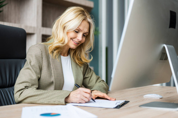 Beautiful mature adult businesswoman with elegant dress sitting at computer desk in the office - Senior woman working at pc online, concepts about business and technology - Fotó, kép