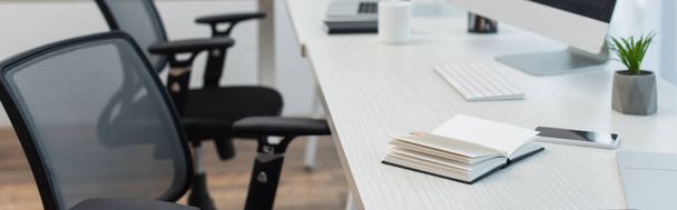 notebook, telefone celular e vaso perto do teclado na mesa perto de cadeiras de escritório borradas, banner - Foto, Imagem