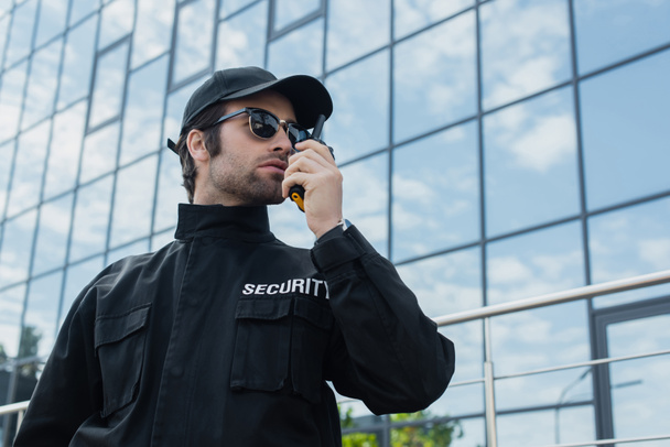 bezpečnostní muž v slunečních brýlích a černé uniformě mluví na rádio nastavit venku - Fotografie, Obrázek