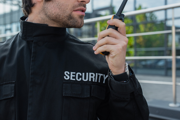 visão parcial de guarda em uniforme com letras de segurança falando em walkie-talkie ao ar livre - Foto, Imagem