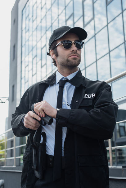 guard in sunglasses and black uniform holding gun and looking away outdoors - Фото, изображение
