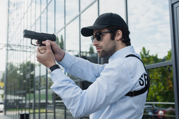 young guard in cap and sunglasses holding gun outdoors - Φωτογραφία, εικόνα