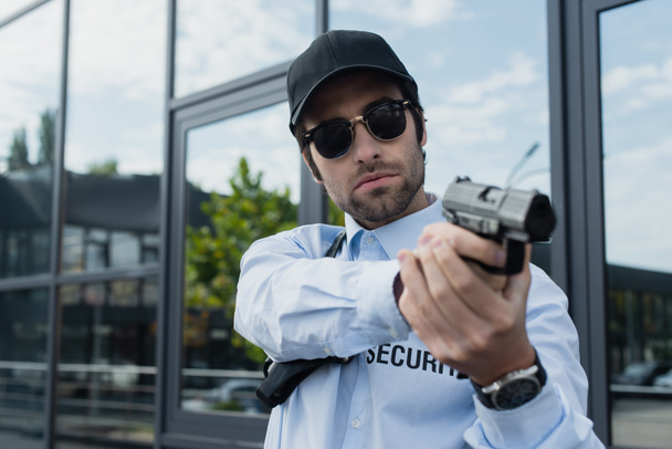 young guard in uniform, black cap and sunglasses standing with gun outdoors - Foto, Imagen