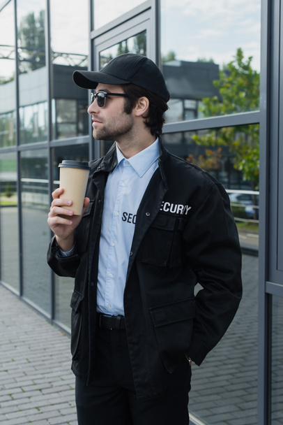 joven hombre de seguridad en uniforme negro y gorra de pie con café en la calle de la ciudad - Foto, Imagen