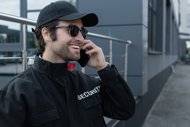 fröhlicher Sicherheitsmann in schwarzer Uniform und Sonnenbrille, der im Freien mit Walkie-Talkie spricht - Foto, Bild