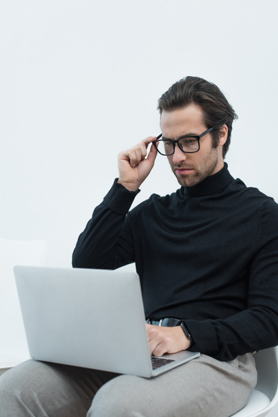 young man in black turtleneck adjusting eyeglasses while working on laptop on grey background - Photo, Image