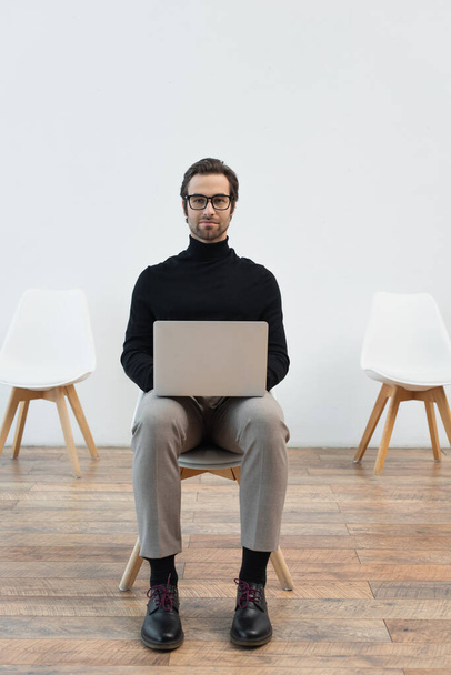 young man in black turtleneck and eyeglasses sitting on chair with laptop and looking at camera - Fotografie, Obrázek