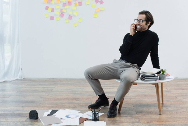 happy man talking on mobile phone while sitting on coffee table near papers on floor - Photo, Image