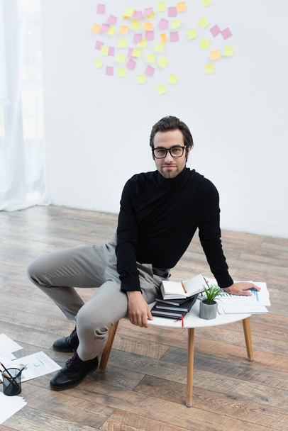 man in black turtleneck and eyeglasses sitting on coffee table near notebooks and looking at camera - Φωτογραφία, εικόνα