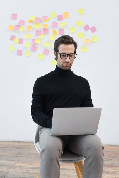 man in black turtleneck and eyeglasses working on laptop near grey wall with sticky notes - Foto, imagen