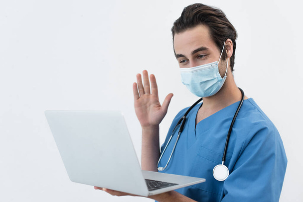 doctor in medical mask waving hand during video call on laptop isolated on grey - Fotó, kép