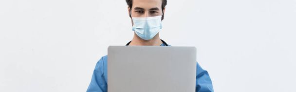 giovane medico in maschera medica utilizzando laptop isolato su grigio, banner - Foto, immagini