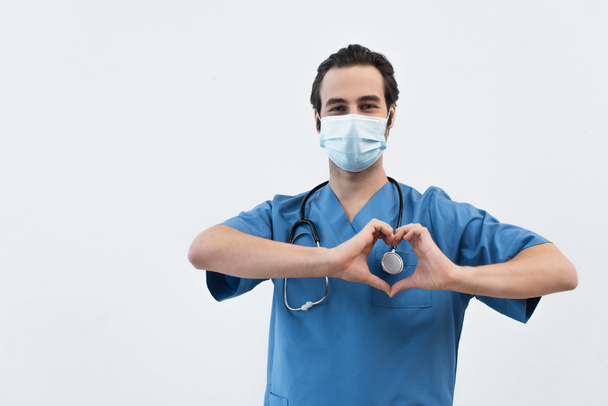 врач в медицинской маске и синей униформе показывает знак сердца с руками, изолированными на сером - Фото, изображение
