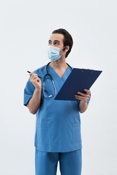 medico in maschera medica distogliendo lo sguardo e indicando con penna isolata su grigio - Foto, immagini
