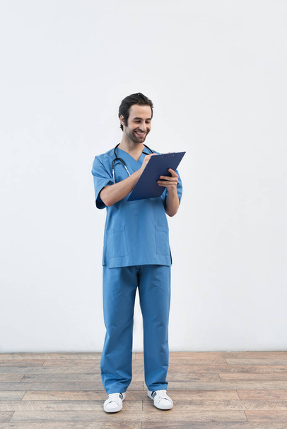 full length view of happy doctor writing on clipboard near grey wall - Foto, Imagem