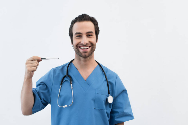 médico alegre en uniforme azul mostrando termómetro electrónico aislado en gris - Foto, Imagen