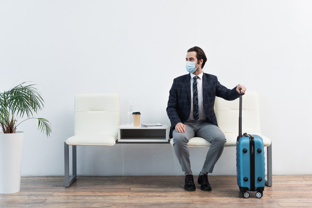 ταξιδιώτης με ιατρική μάσκα κάθεται στο σαλόνι αναχώρησης κοντά στη βαλίτσα και κοιτάζοντας μακριά - Φωτογραφία, εικόνα