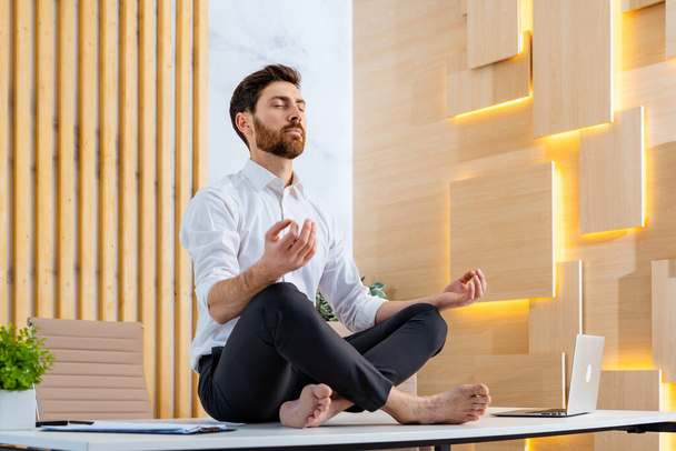 Businessman with elegant suit sitting at computer desk in the office meditating - Adult man doing yoga meditation and deep breathing at work to calm and relax the soul - Foto, imagen
