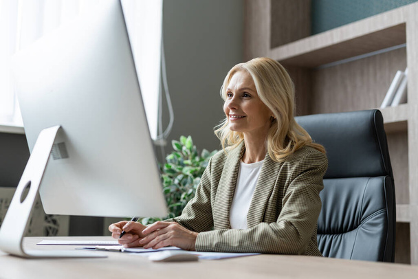 Beautiful mature adult businesswoman with elegant dress sitting at computer desk in the office - Senior woman working at pc online, concepts about business and technology - Foto, Imagen