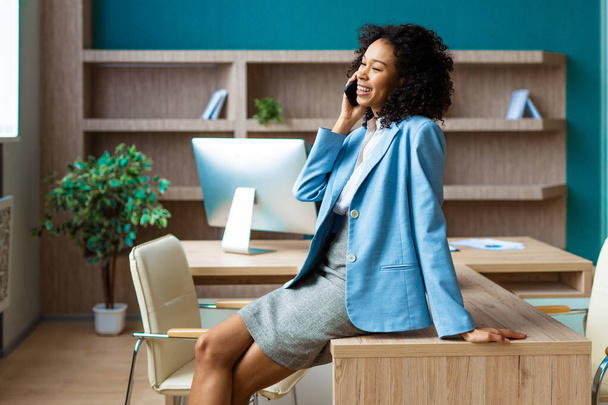 Beautiful adult businesswoman with elegant dress standing in a business studio and using a mobile phone app - African american woman portrait in a business meeting, concepts about business and technology - Photo, image