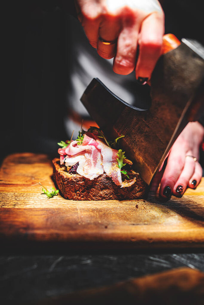 Woman chef hand cutting tasty sandwich with axe on wooden plate - 写真・画像