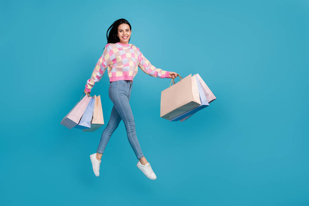 Photo of cute charming woman wear pink sweater jumping high holding bags empty space isolated blue color background - Фото, изображение