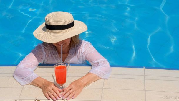 ストローハットで金髪の女性を笑顔にし、晴れた夏の日にスイミングプールの近くでオレンジカクテルを飲みます。夏の気分の概念、休暇、すべてが含まれる - 写真・画像