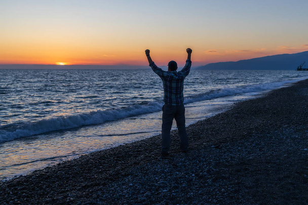 Silhouette eines Mannes, der sich abends am Meer an der Küste freut. Hände erhoben bei Sonnenuntergang - Foto, Bild