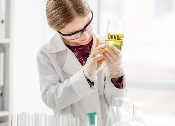 Smart girl doing scientific chemistry experiment wearing protection glasses, holding bottle with title danger. Schoolgirl with chemical equipment on school lesson - Zdjęcie, obraz