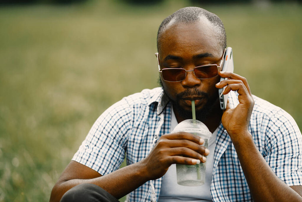 Black american man in a sunglasses sitting on a grass and talking the phone - 写真・画像