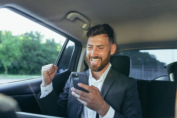 Mutlu ve başarılı erkek işadamı araba yolcusu haberleri telefondan okur, zaferden memnun kalır ve haberleri alır. - Fotoğraf, Görsel