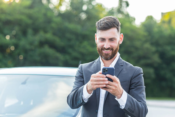 Cheerful and successful male businessman car salesman uses the phone near the parking lot to sell cars - Foto, afbeelding