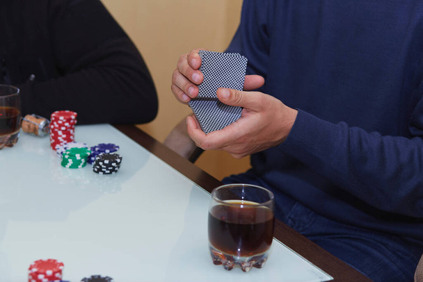 Τα χέρια του ανθρώπου ανακατεύουν τα χαρτιά σε ένα παιχνίδι πόκερ. Πατατάκια, κάρτες, ένα ποτήρι ουίσκι στο τραπέζι με αντανάκλαση. Λέσχη πόκερ. - Φωτογραφία, εικόνα