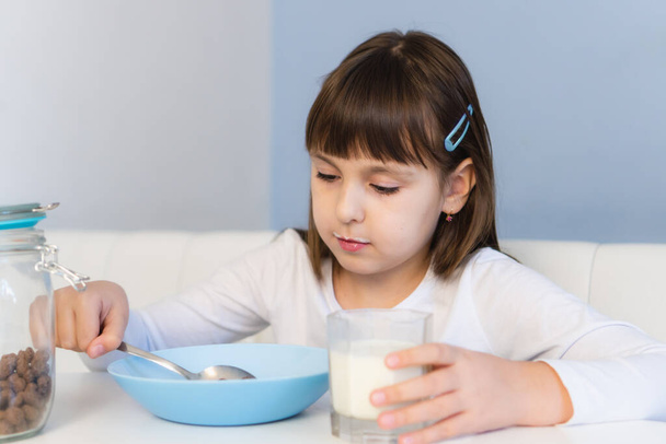 Menina come bolas de milho de chocolate com leite sentado à mesa branca. Criança tomar café da manhã - Foto, Imagem
