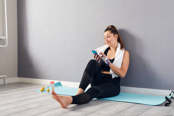 Tired Fitness woman wearing black tracksuit sitting making selfie shot on mobile phone after training. Girl live streaming home workout, giving fitness class online during self quarantine. - Photo, image