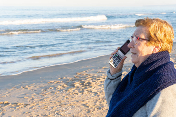 Пожилая женщина на пляже у моря, разговаривает по телефону адаптированному для пожилых людей - Фото, изображение