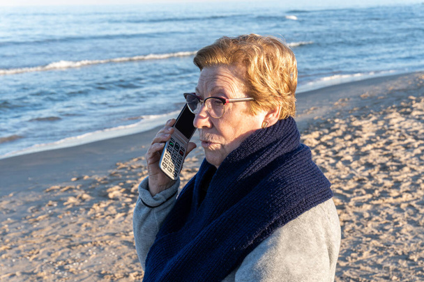 Mujer mayor en la playa cerca del mar, hablando por teléfono adaptado para personas mayores - Foto, imagen