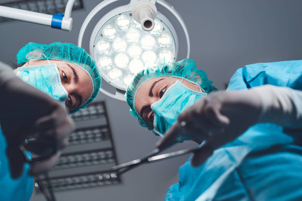 Desde abajo cirujanas en uniforme médico usando herramientas profesionales mientras están de pie bajo luz brillante en quirófano - Foto, imagen