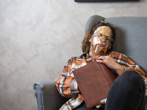 A slender brunette is reading a book with a gold leaf mask on her face. relaxing with beauty treatments at home. - Photo, Image