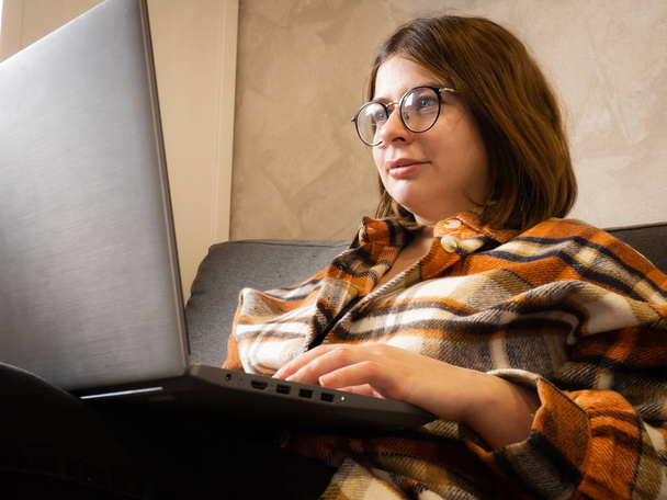 νεαρή κοπέλα με γυαλιά που εργάζεται σε ένα φορητό υπολογιστή κάθεται στο σπίτι στο κρεβάτι. - Φωτογραφία, εικόνα