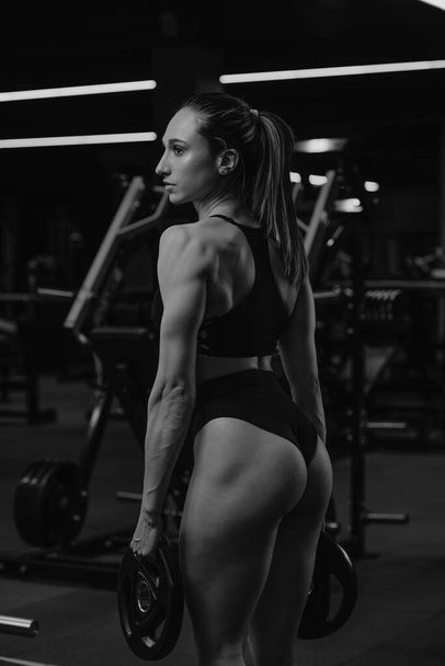 Muskularna kobieta z plecami pozuje na siłowni z dwoma gumowymi ciężarkami. Dziewczyna w koszulce i sportowej bieliźnie ćwiczy z żelaznymi talerzami. - Zdjęcie, obraz