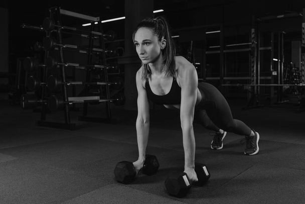 Μια μυώδης γυναίκα είναι push ups με αλτήρες σε ένα γυμναστήριο. Ένα κατάλληλο κορίτσι σε ένα μπλουζάκι και σπορ κολάν κάνει μια προπόνηση στο στήθος. - Φωτογραφία, εικόνα