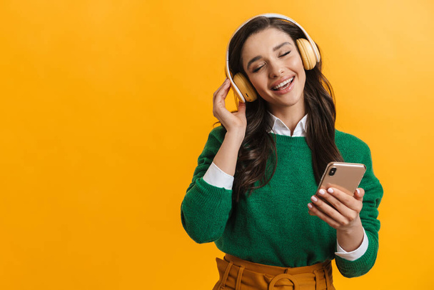 Portrait of a happy smiling casual woman standing over yellow background listening to music with headphones holding mobile phone - Fotografie, Obrázek